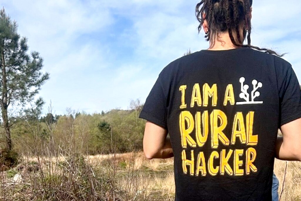 Persona de espaldas en un campo, con camiseta negra que dice «I AM A RURAL HACKER» en letras amarillas y un pequeño icono de planta