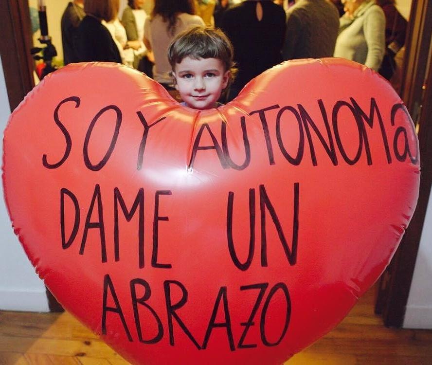 Niña asomada tras un globo corazón rojo con el mensaje «Soy autónom@ dame un abrazo» en un encuentro de comunidad
