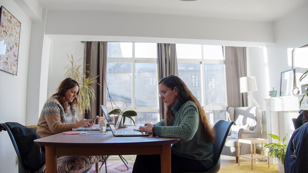 Dos mujeres trabajando con portátiles en la mesa del salón, con luz natural y ventanales