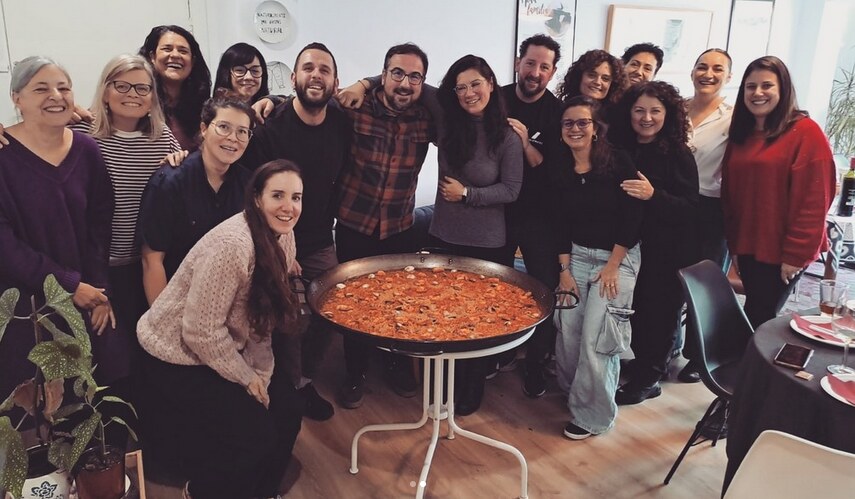 Un grupo diverso de personas sonrientes posando juntas alrededor de una gran paella tradicional en un ambiente comunitario y acogedor