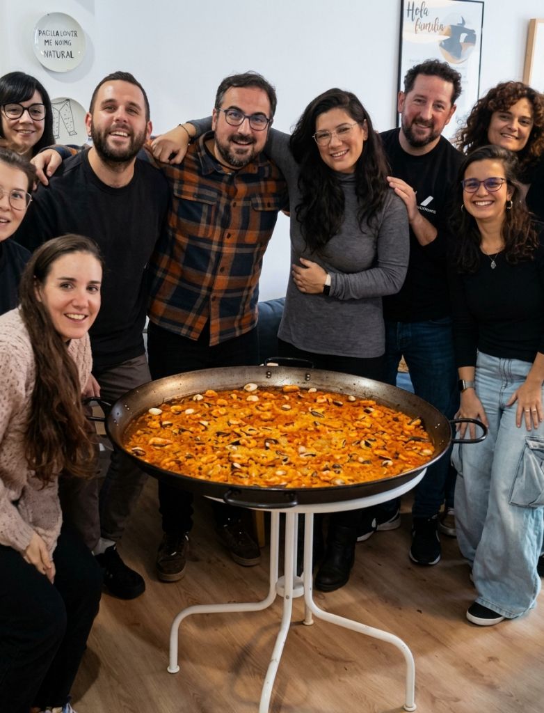 Un grupo diverso de personas sonrientes posando juntas alrededor de una gran paella tradicional en un ambiente comunitario y acogedor