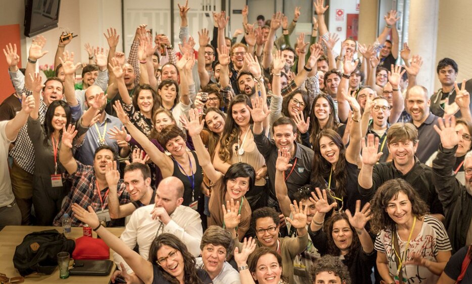 Gran grupo de asistentes de la Coworking Spain Conference con las manos en alto, sonriendo en un espacio de coworking junto a la puerta de cocina