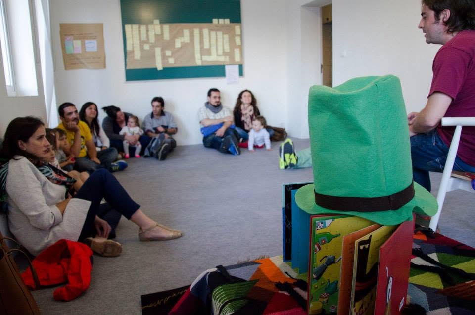 Pablo Cañiza (cuenta cuentos) en el suelo con sombrero verde, libros infantiles y muro de los deseos al fondo