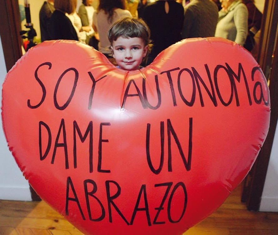 Niña asomada tras un globo corazón rojo con el mensaje «Soy autónom@ dame un abrazo» en un encuentro de comunidad