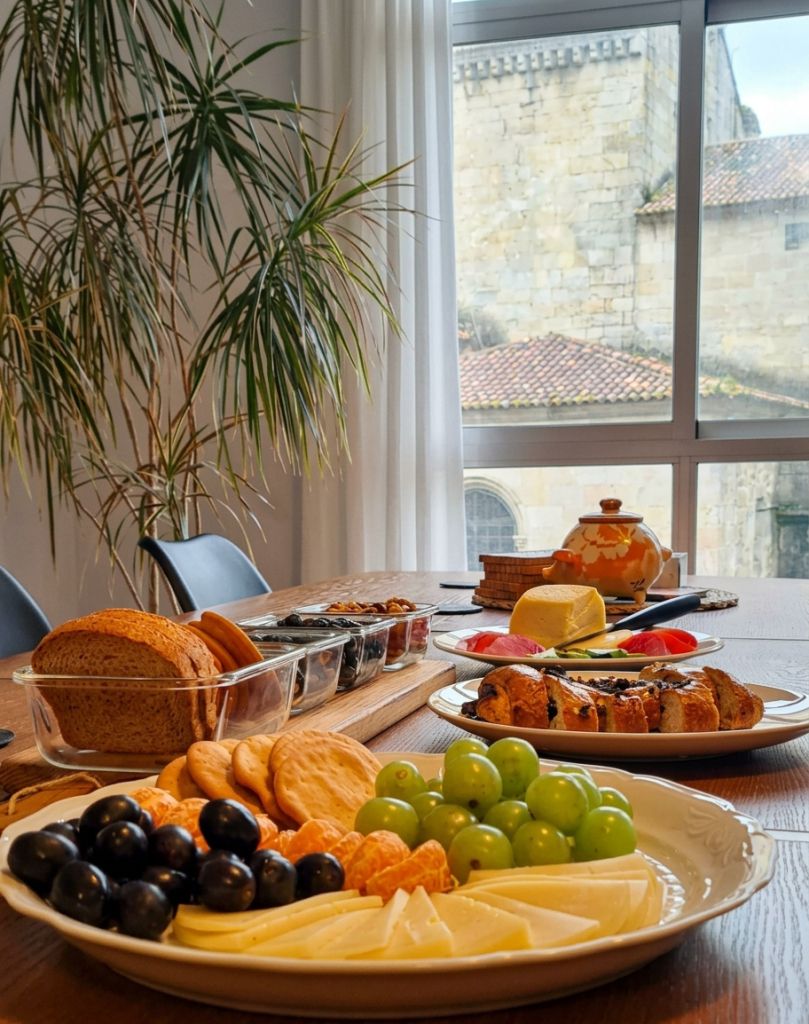 Bodegón de aperitivos sobre mesa de madera: uvas, naranja, queso y pan frente al ventanal