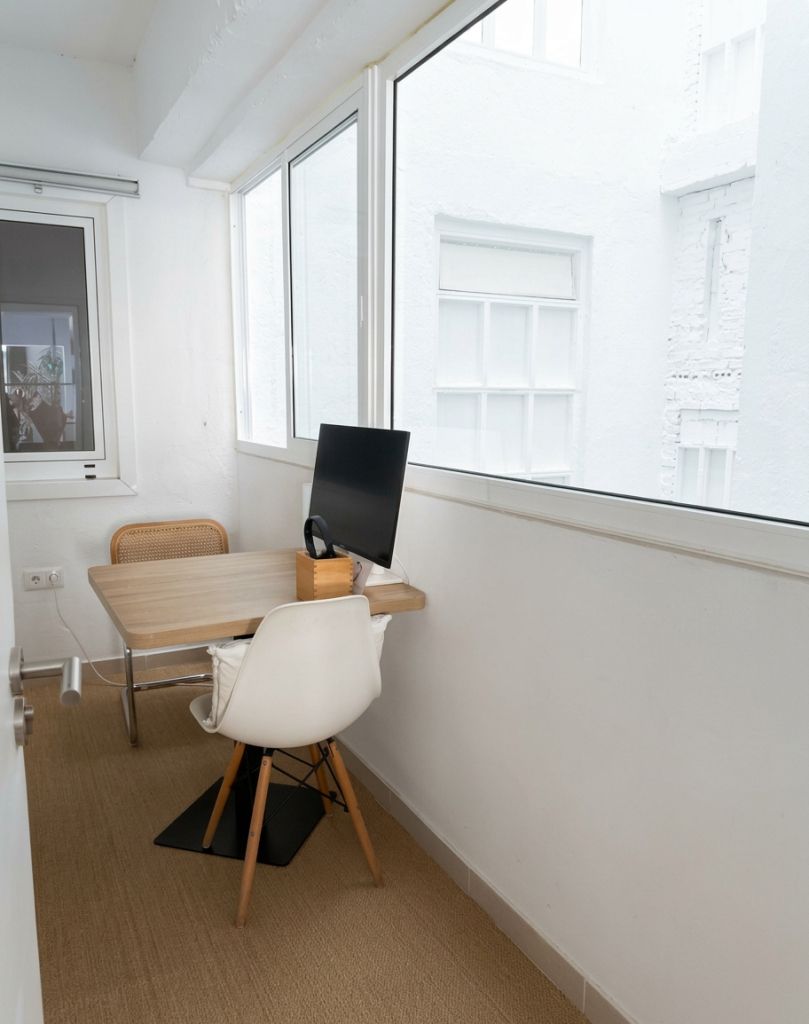 Sala de reuniones pequeña con mesa de madera, monitor, silla blanca y ventanales al patio interior
